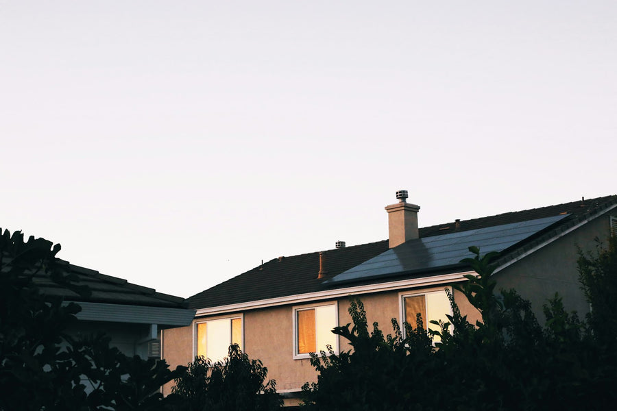 A two-story stucco house at dusk with warm light in two upstairs windows, solar panels on the roof and trees in the foreground.