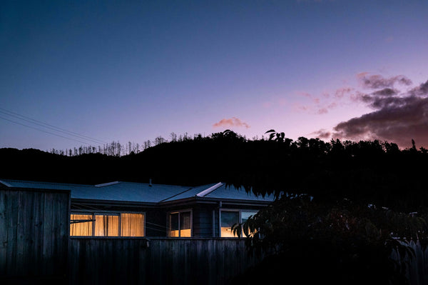 A single-story house at dusk with lit windows behind a wooden fence, the sky turning purple