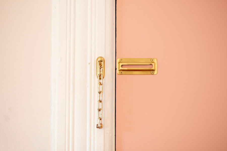 A brass door chain mounted on a white door frame next to a brass strike plate on a peach-painted door.