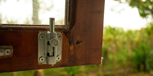 Close-up of a metal barrel-bolt latch screwed to a dark wooden window frame, with green foliage soft behind the glass.