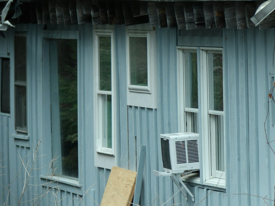 A white window air-conditioning unit sitting in the lower sash of a double-hung window on a blue-grey wooden house, held up by a metal support bracket.